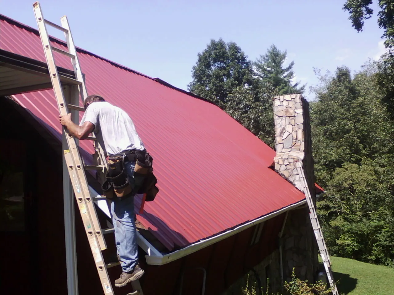Roofer on a ladder beside a red metal roof and stone chimney — Soffit & Fascia Repair by Roofing Brothers in Moss Point